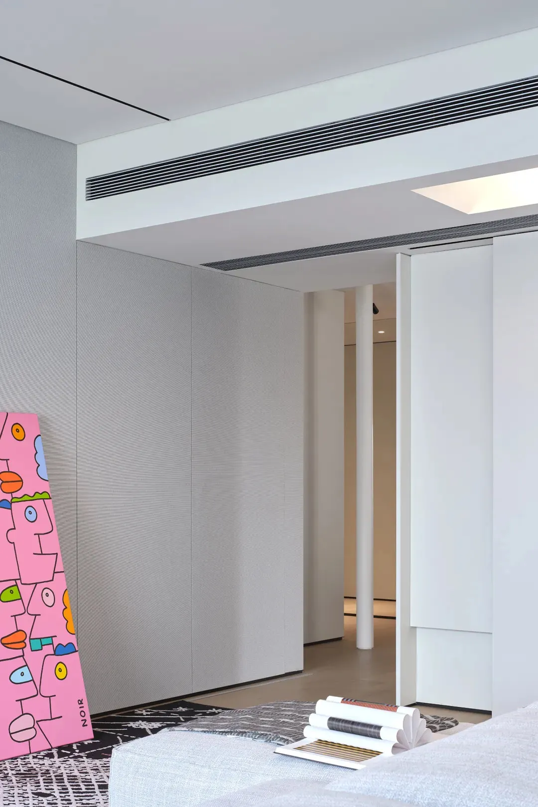 A modern living room featuring minimalist white paneling, with a pink artwork leaning against the wall and a stack of books o