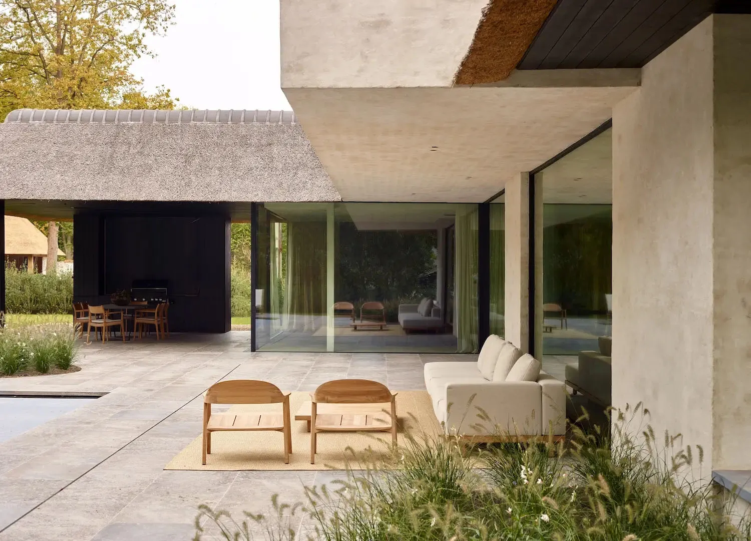 A minimalist residential exterior featuring concrete walls, large glass windows, and a thatched-roof pavilion.