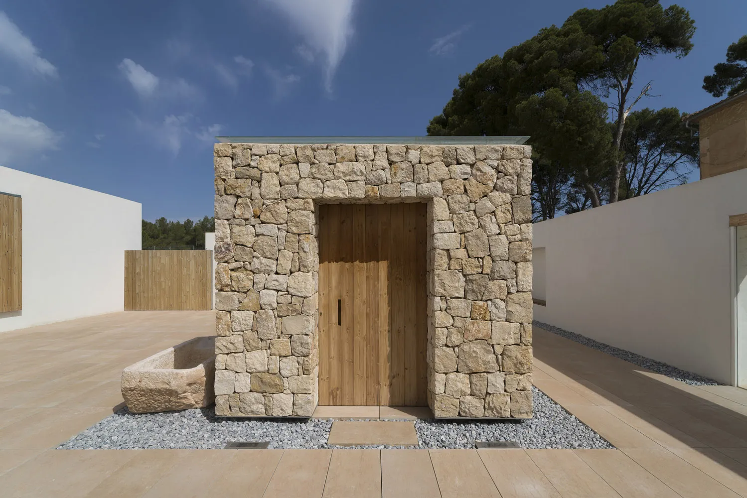 A minimalist stone building exterior with a wooden door and gravel pathway.