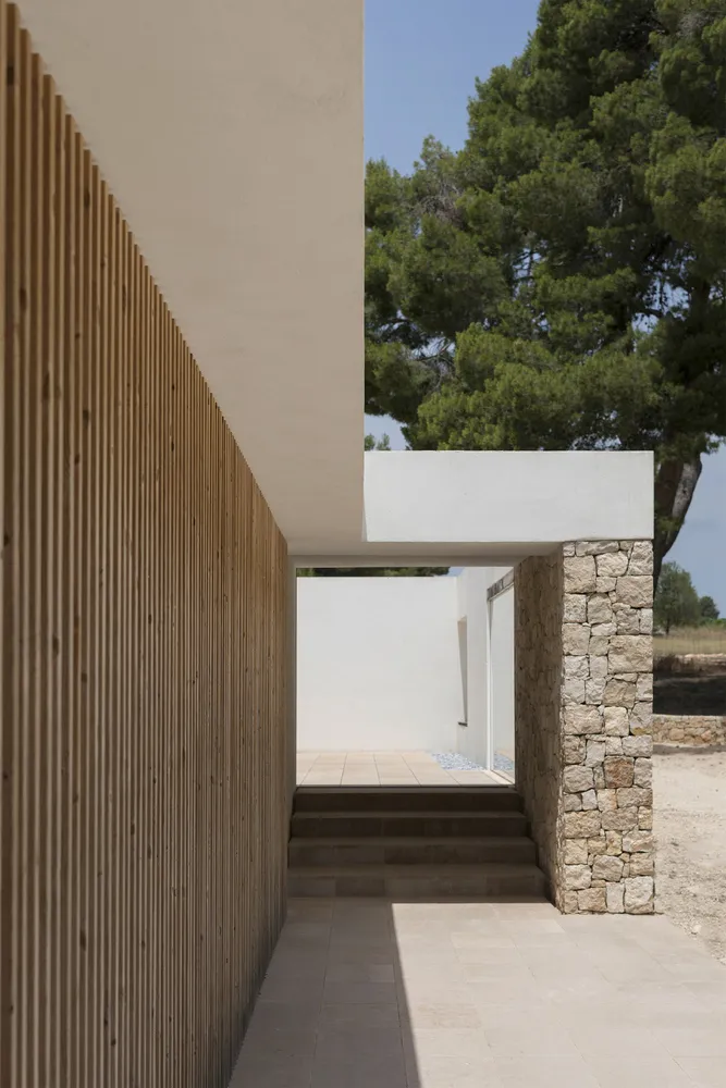 Exterior view of a modern farmhouse featuring a wooden fence, concrete columns, and stone steps leading up to a white wall.