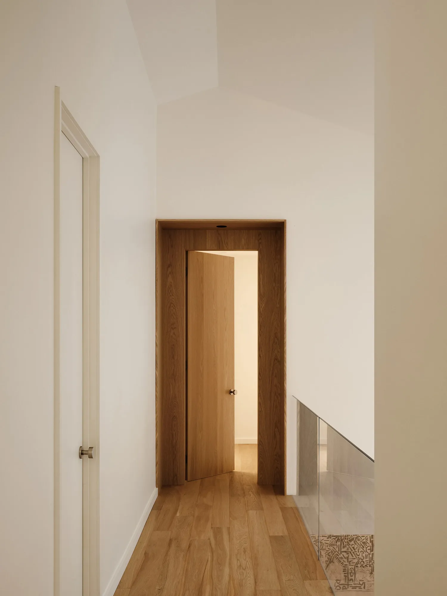 A minimalistic architectural space featuring a wooden door framed by glass, accentuated by a wooden floor and white walls.