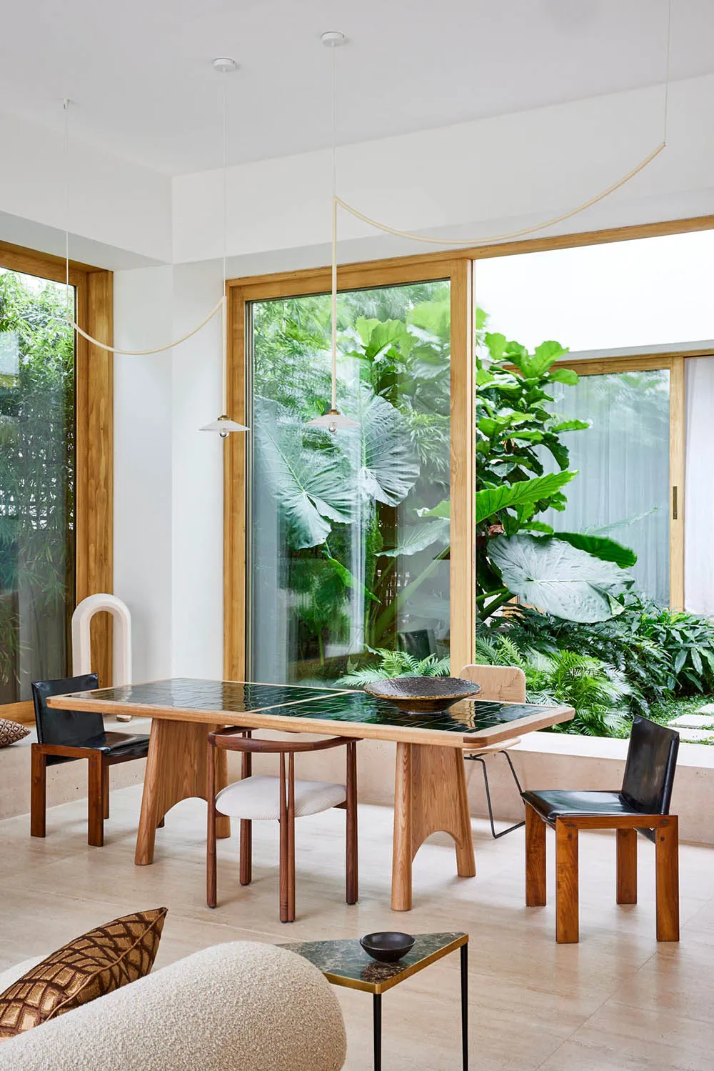 A modern dining room featuring a wooden table with green tiles, surrounded by natural light and lush greenery outside the win