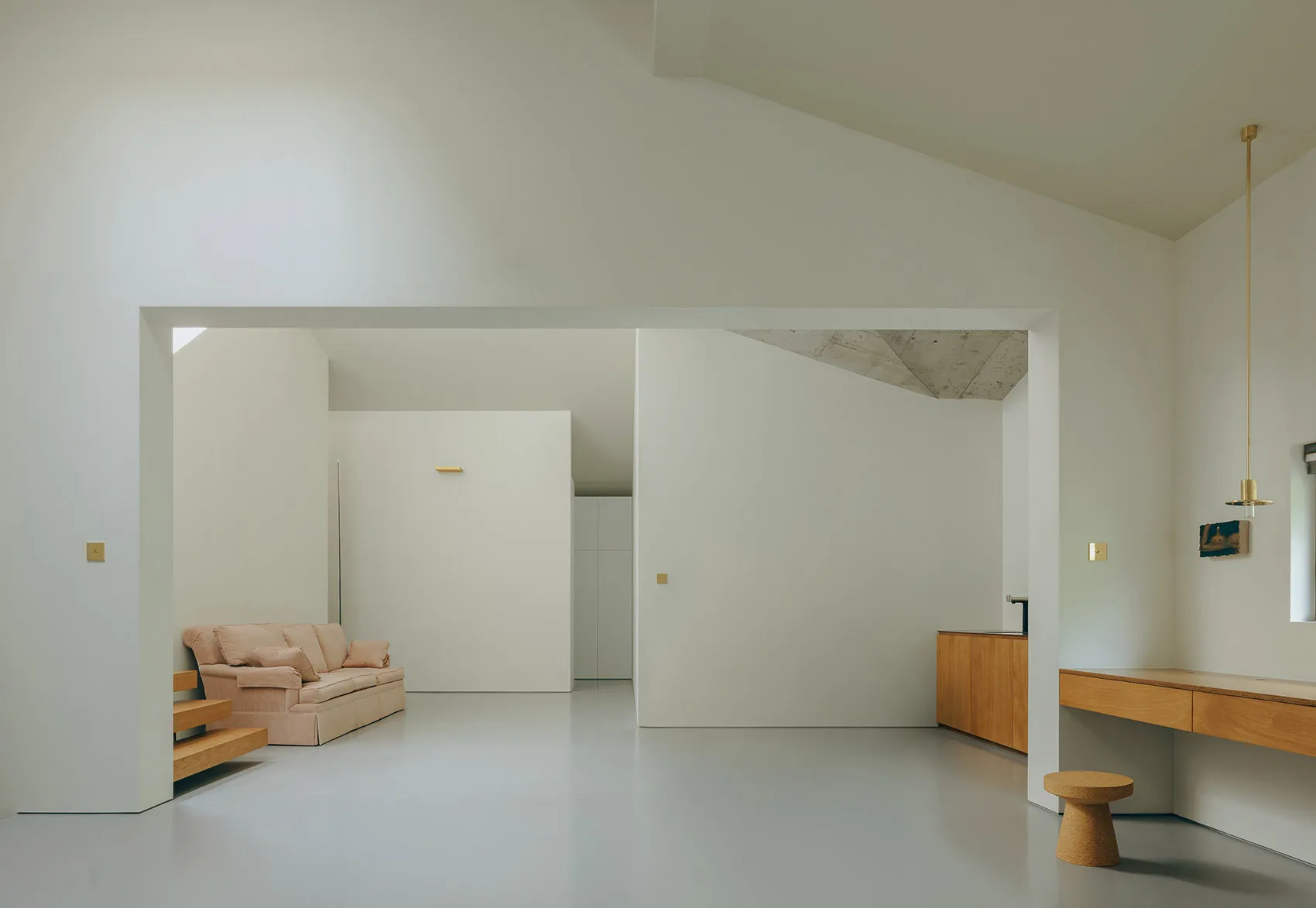 A minimalist living room with concrete and white walls, a low wooden bench, and a beige sofa with pillows.