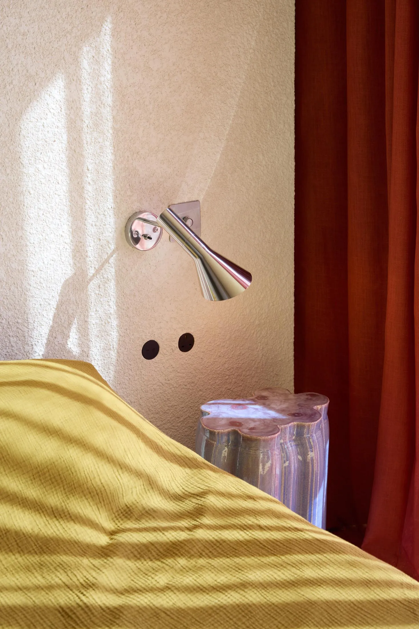 A modern bedroom featuring a bright, metallic wall lamp, yellow bedding, and a textured wall with sunlight shining through th