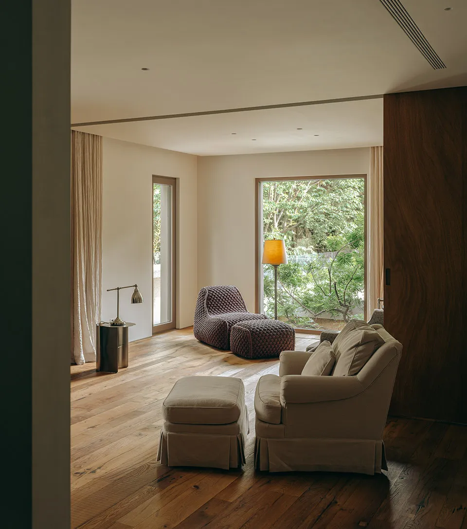 A modern living room with wood flooring, a beige sofa, and a floor lamp next to a window with trees outside.