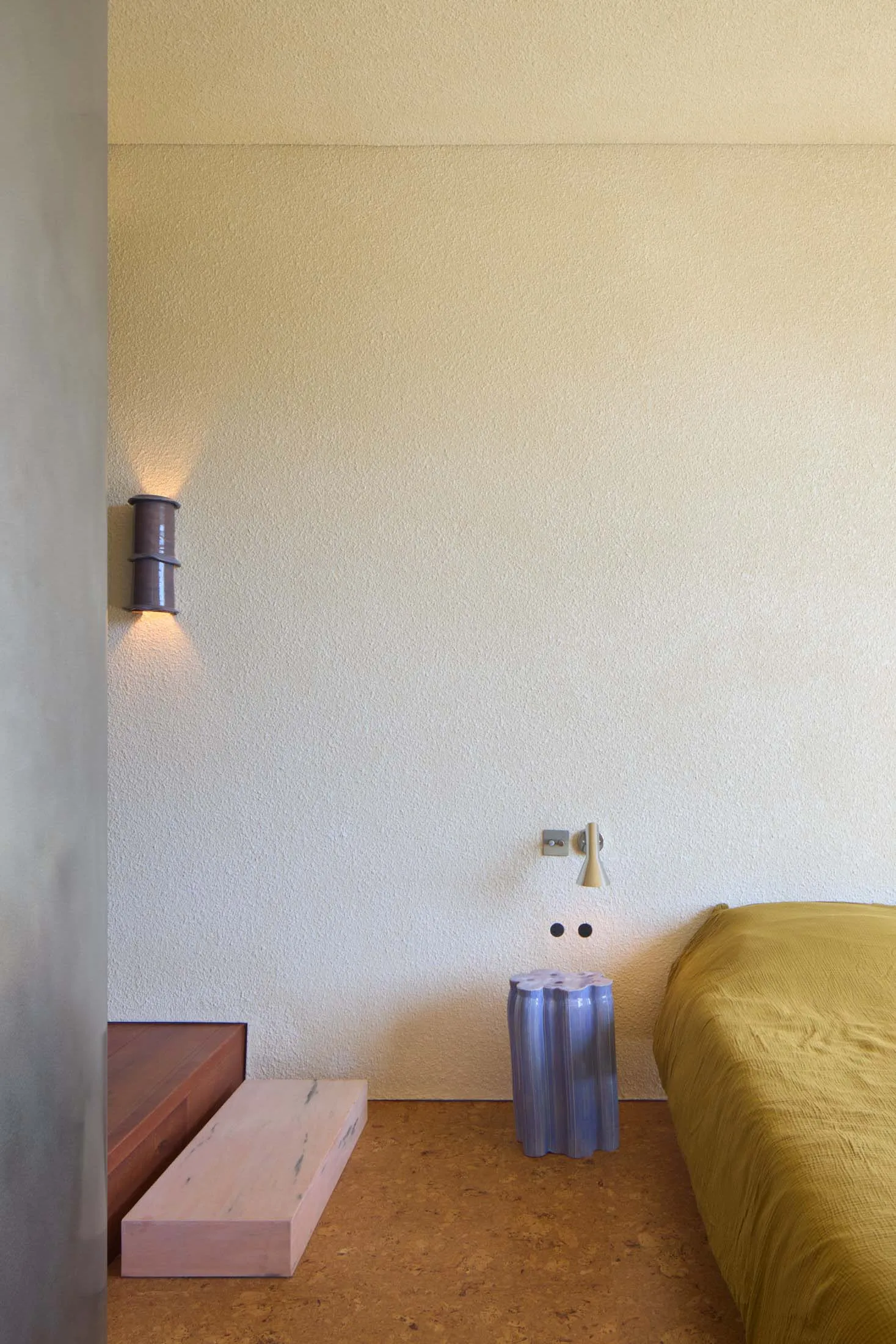 A minimalist bedroom featuring a concrete wall, a yellow bedspread, and a pink wooden bench.