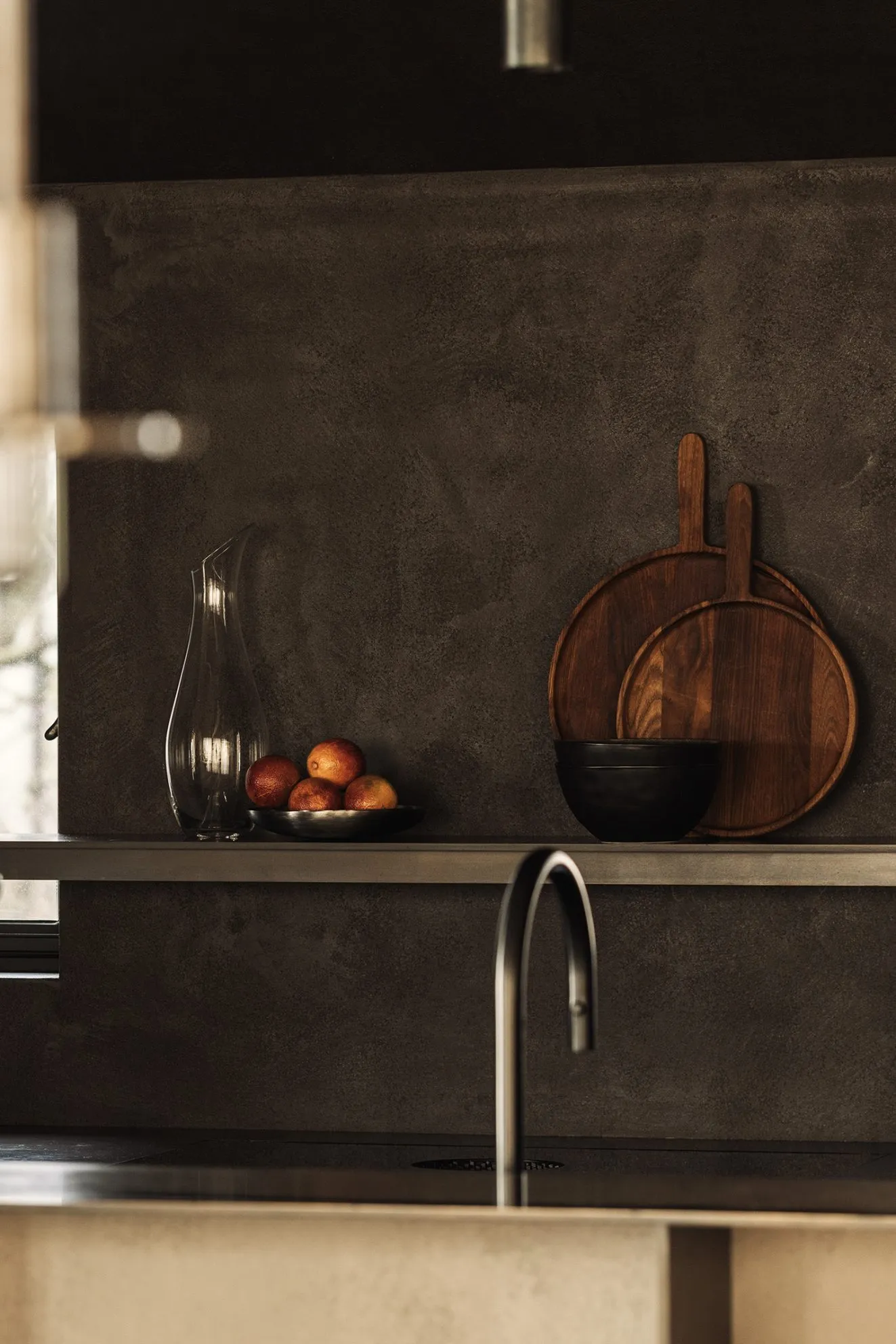 Minimalist kitchen with a concrete backsplash, featuring a stainless steel faucet, a bowl of oranges, and a carafe.
