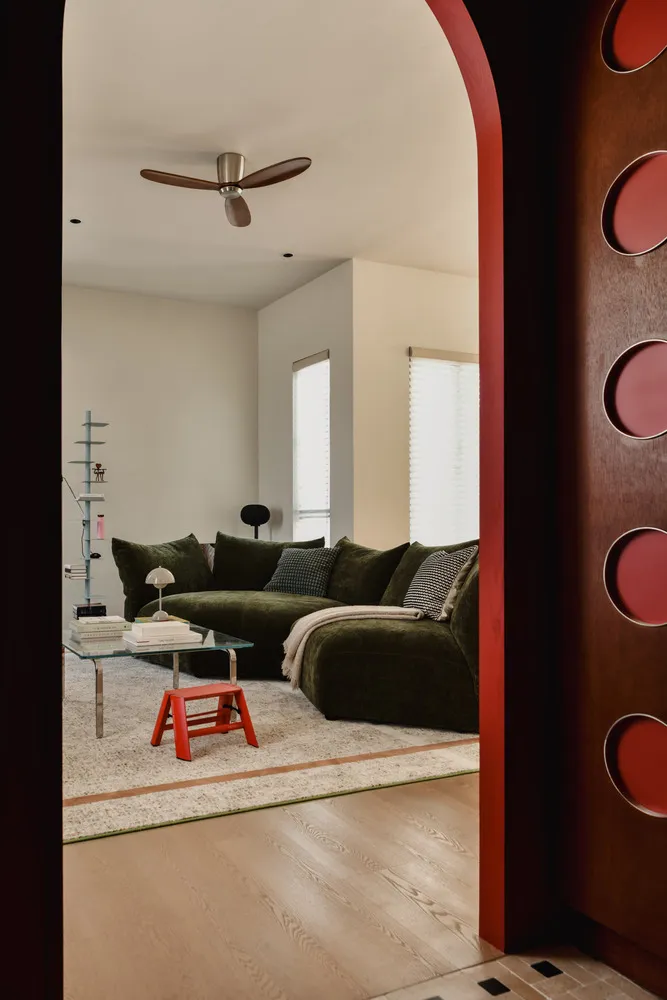 Modern living room featuring a green sectional couch, glass coffee table, and red stool on a wooden floor.