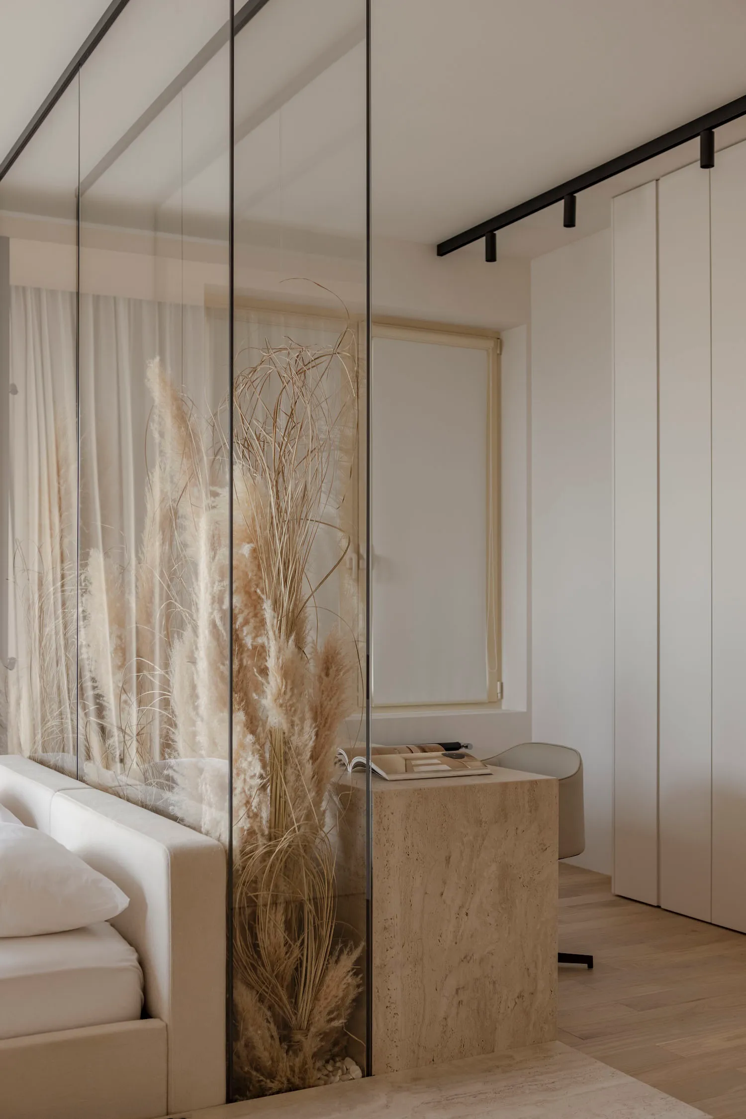 A modern minimalist bedroom featuring a wooden bed frame, glass closet doors, and fabric curtains with pampas grass plant.