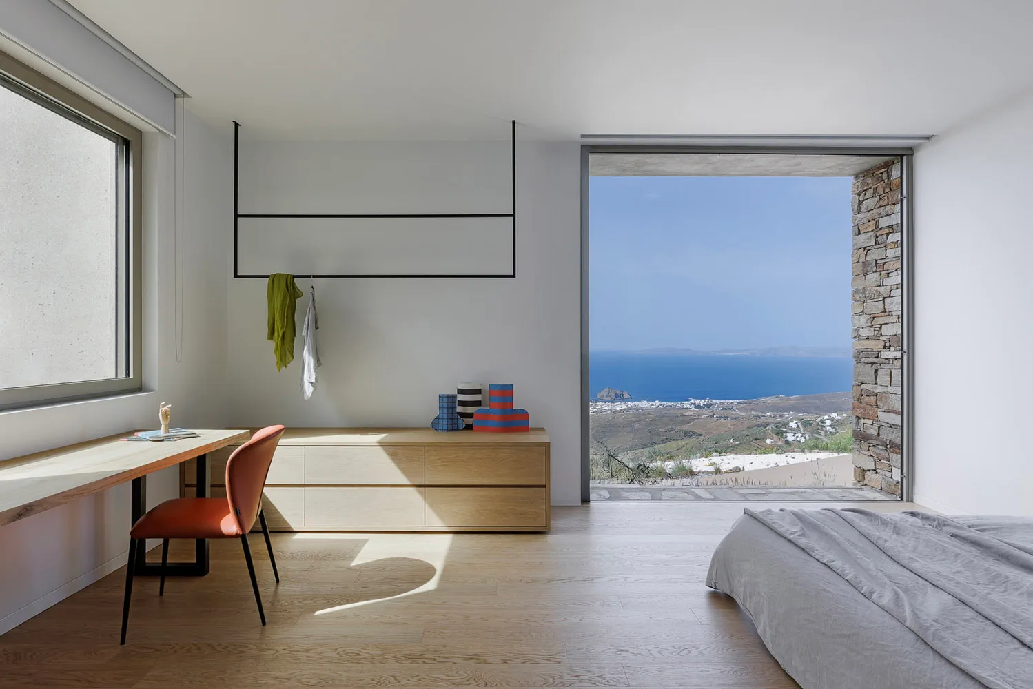 A minimalist bedroom with a wooden floor, white walls, and a large window overlooking the ocean.