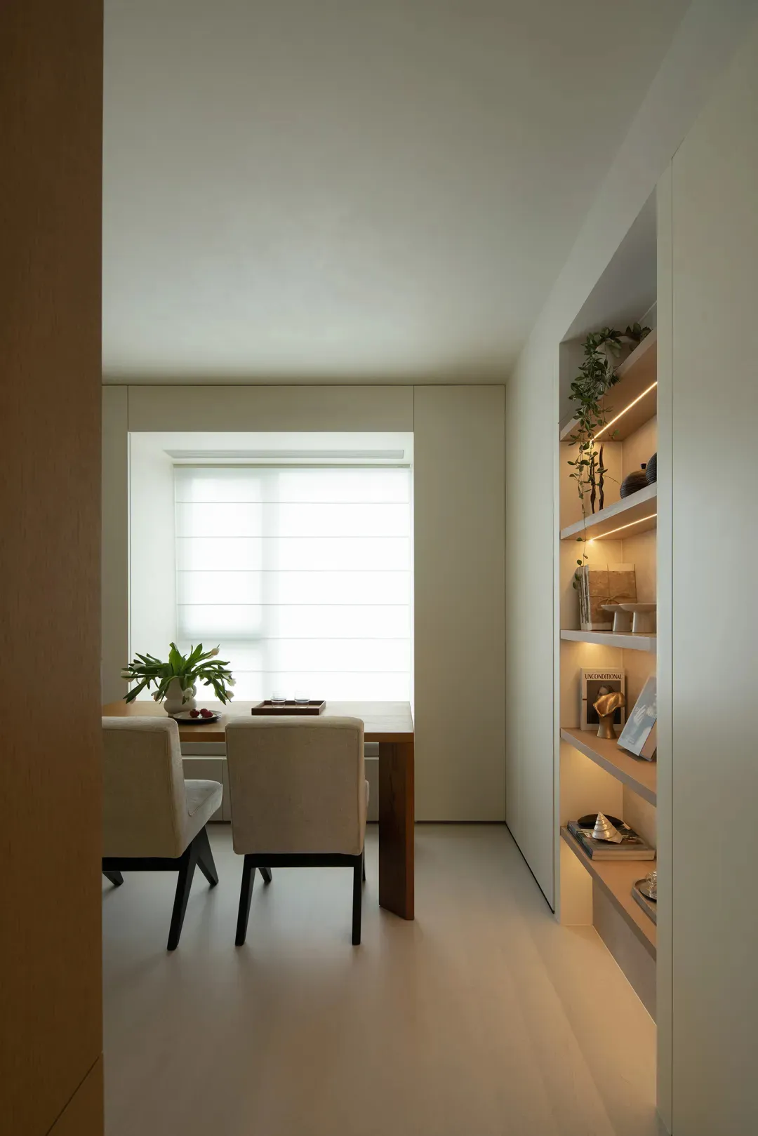 A minimalist dining room featuring a wooden table and beige chairs against a white wall with a large window providing natural