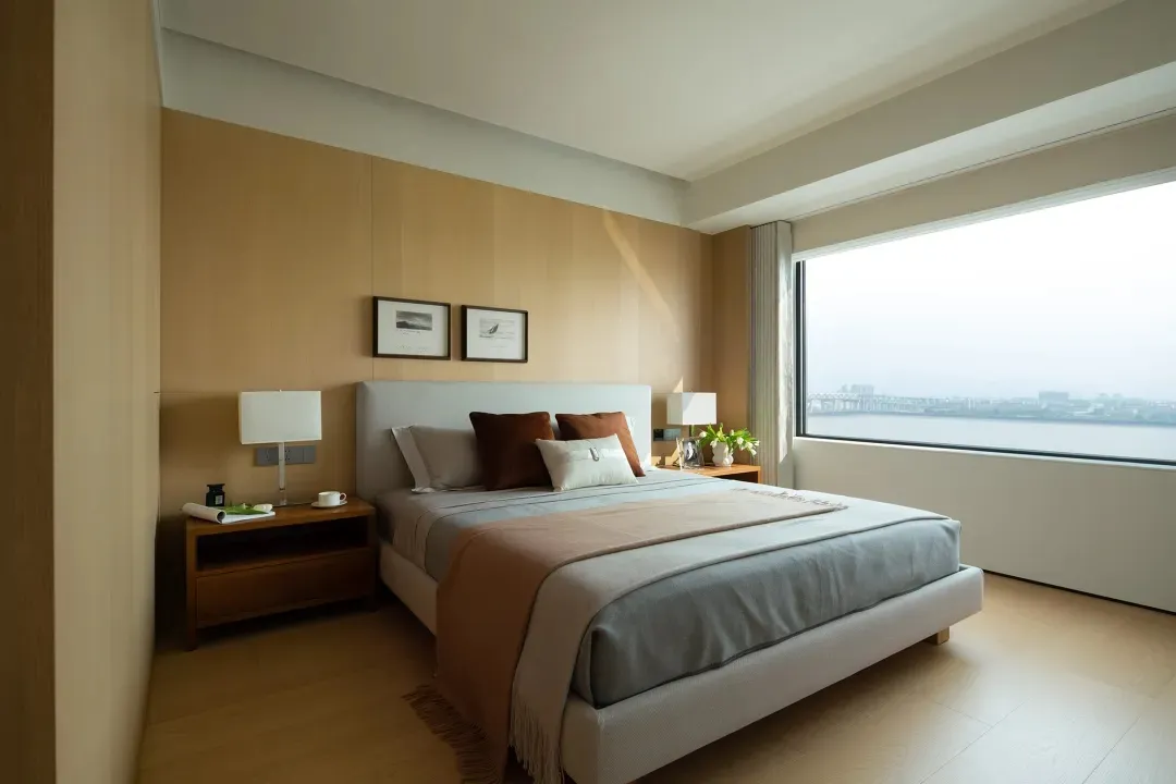 A modern, minimalist bedroom featuring a large window with a view of the city, a white bed with neutral-colored bedding and t