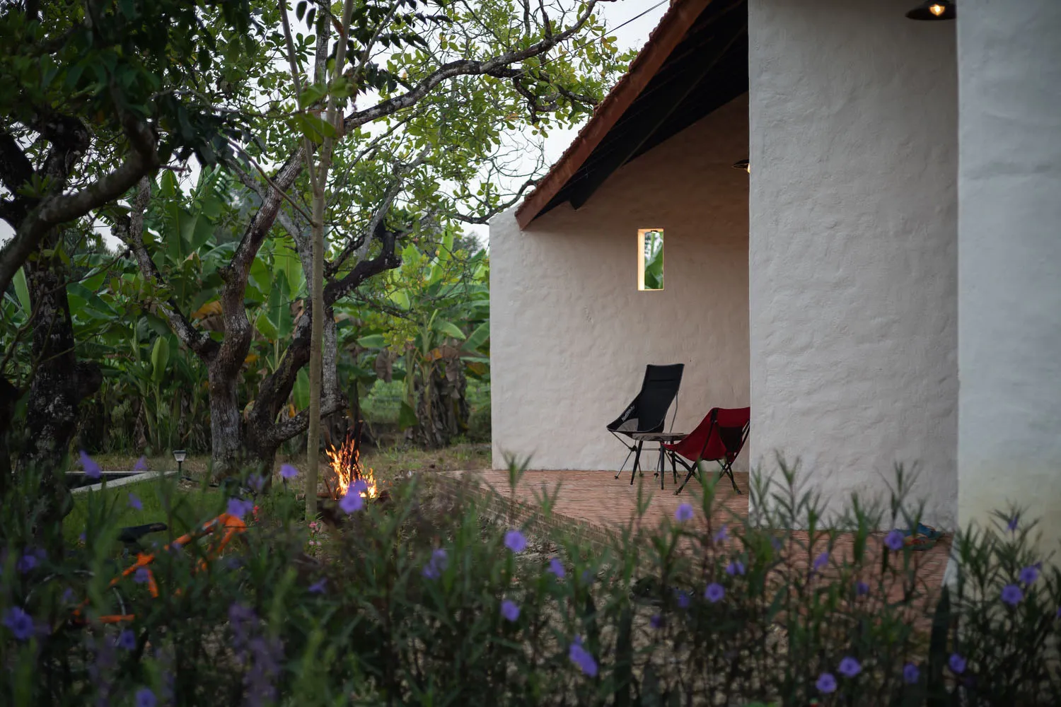 Outdoor patio with concrete floor, chairs, and a fire pit surrounded by greenery and flowers.