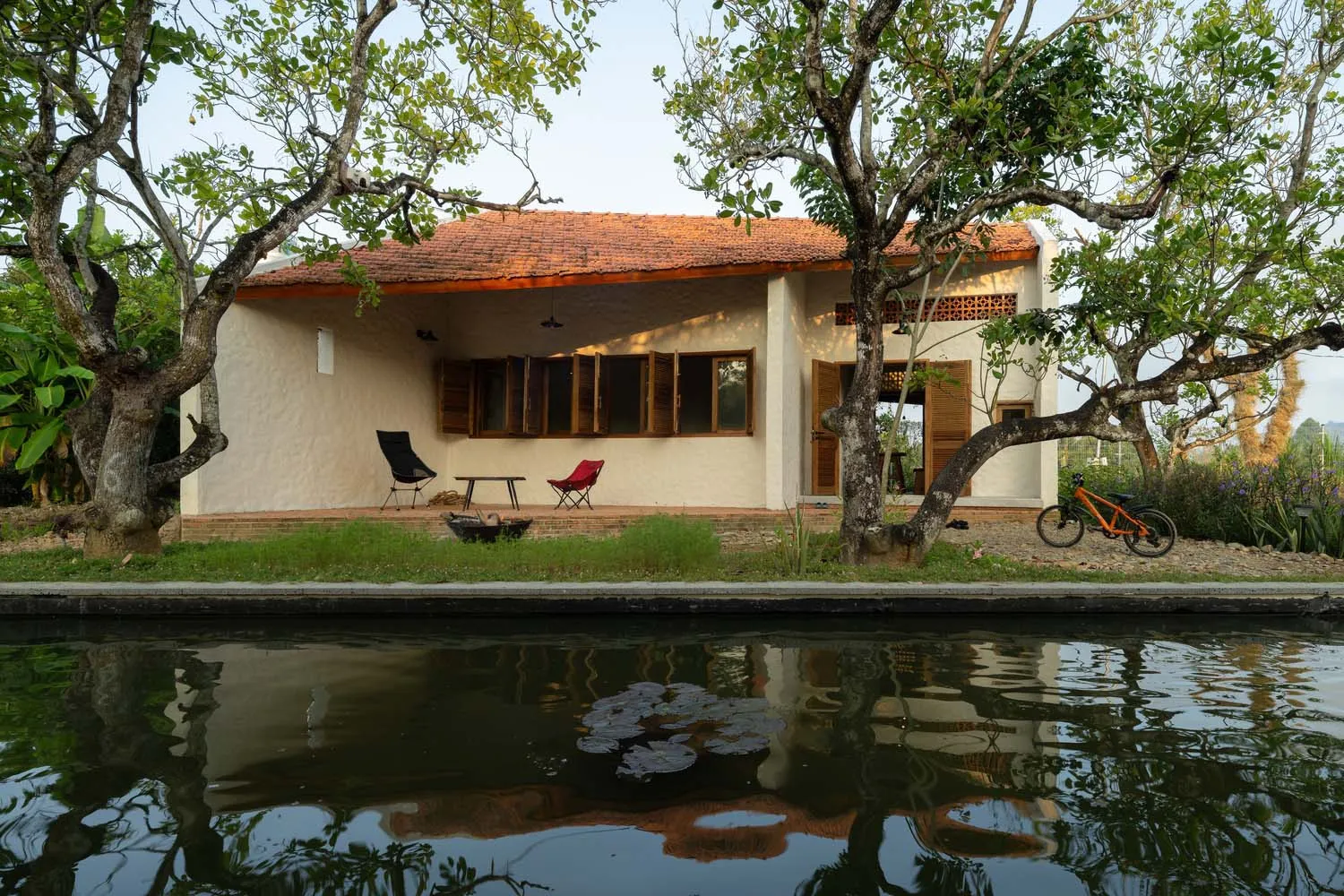 Traditional village house with red tiled roof, white walls, and brick exterior by a calm water body.