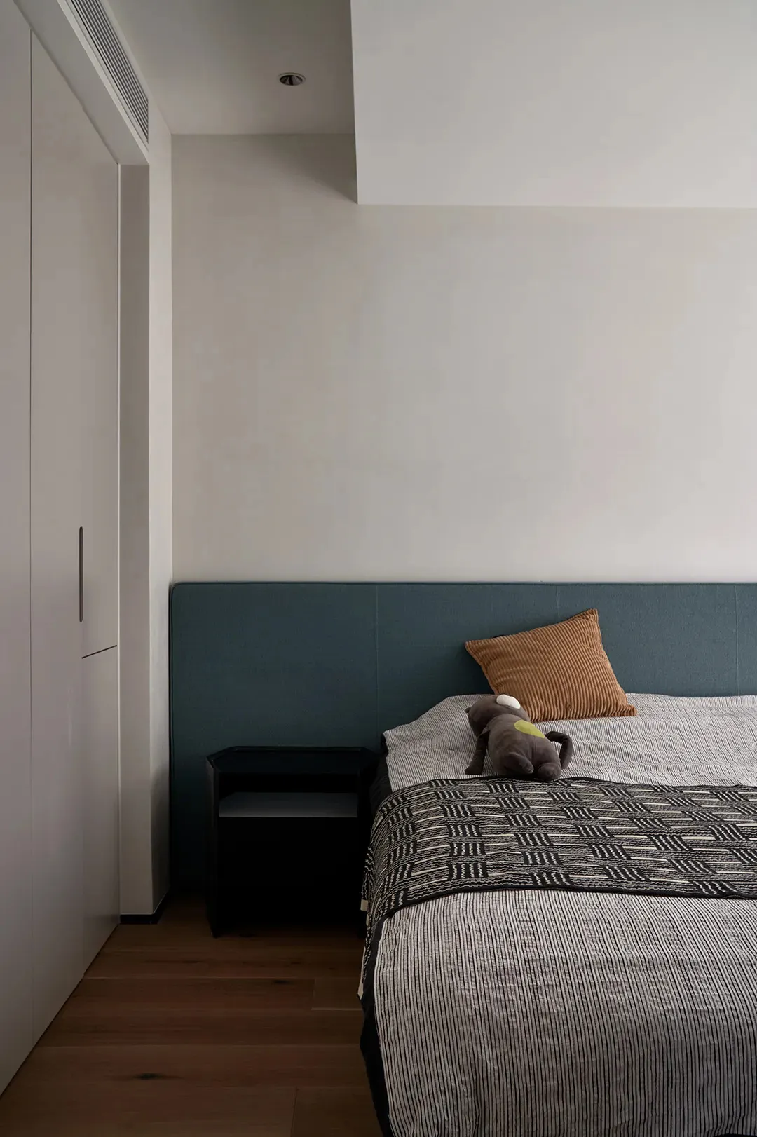A minimalist bedroom featuring a fabric-covered bed with a wooden headboard, a small black side table, and a brown plush toy