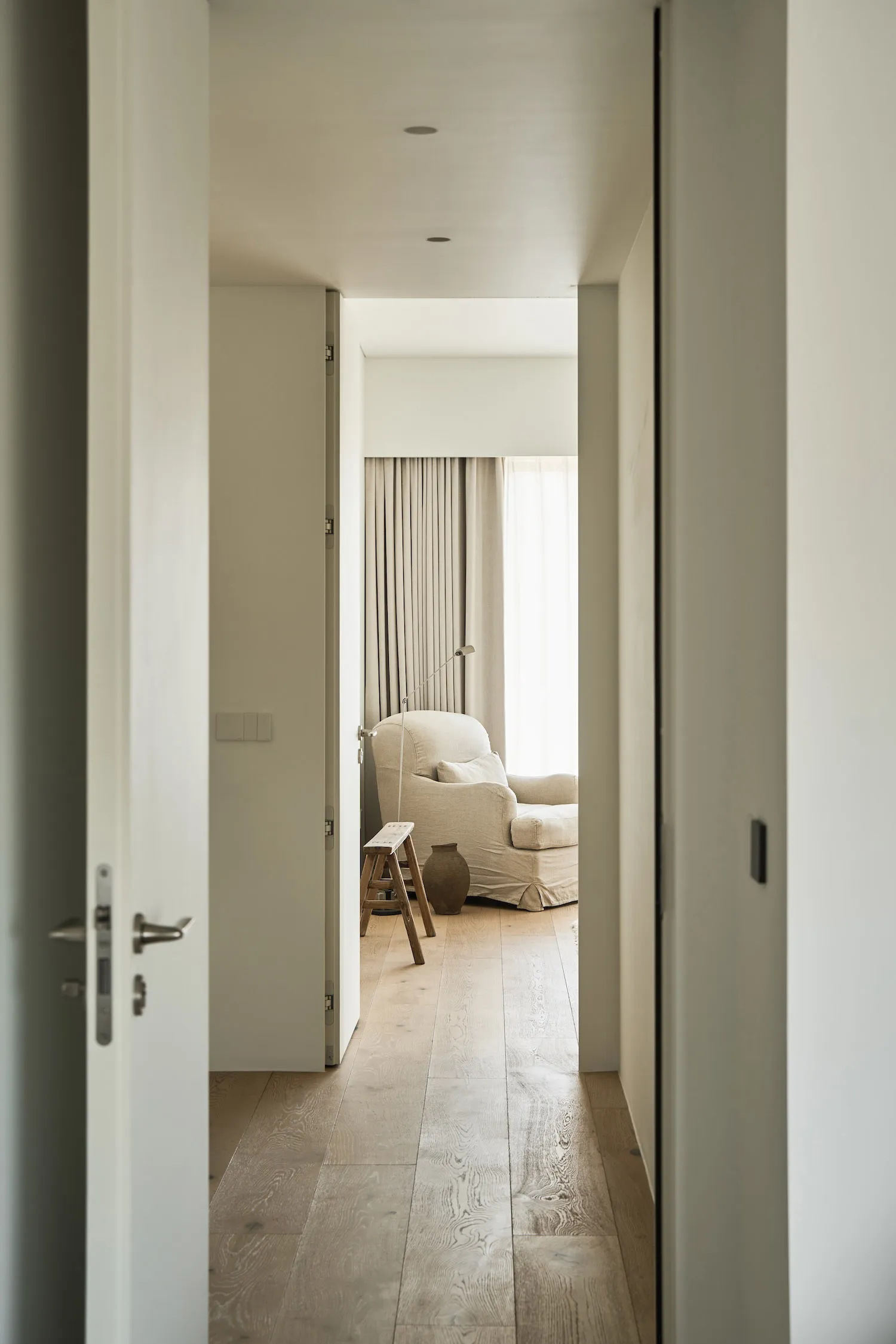 A minimalist residential room with a wooden floor, a beige armchair, and a small wooden stool.