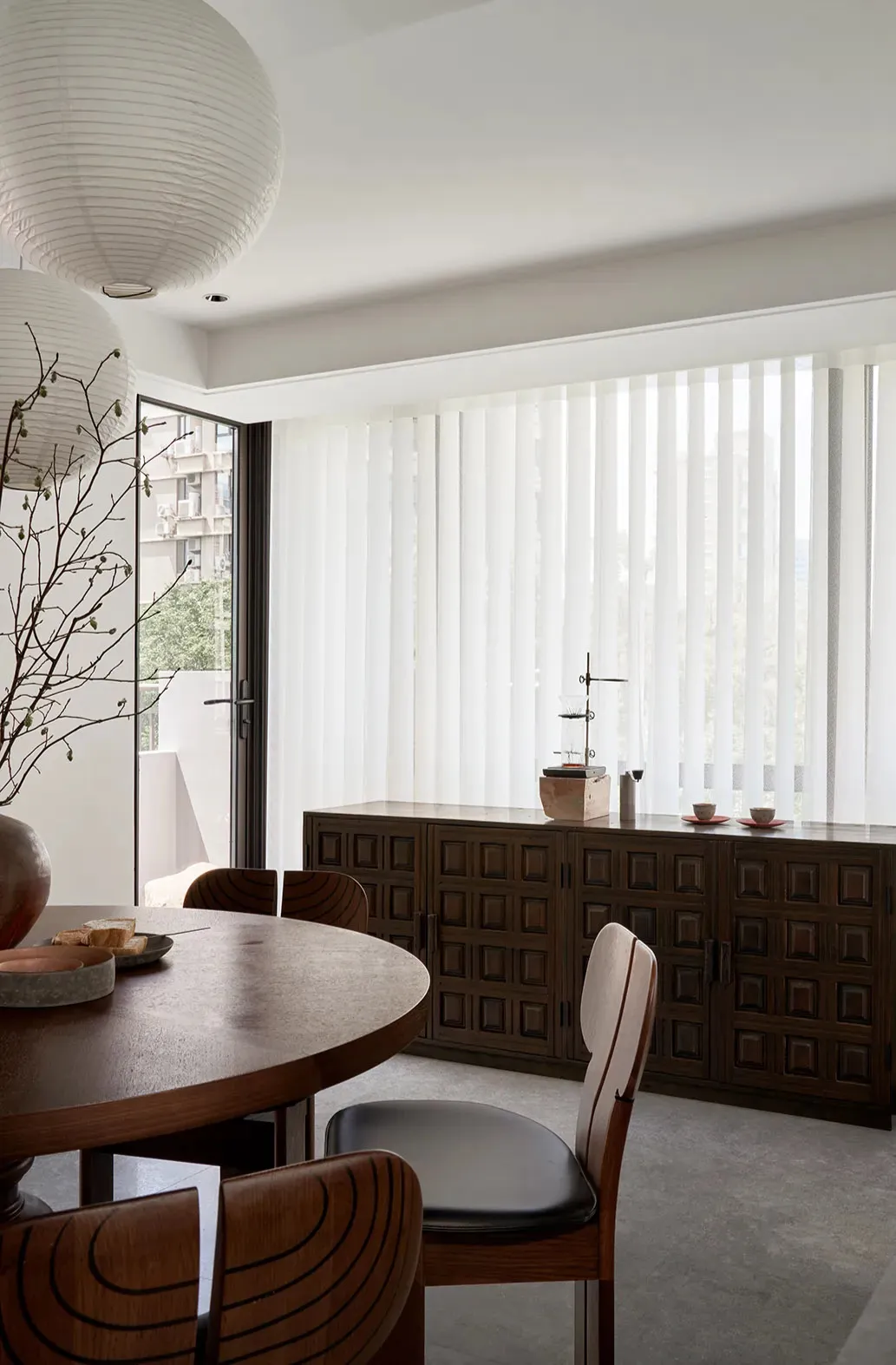 Modern dining room with round wooden table, black chairs, wooden cabinet, and white sheer curtains.