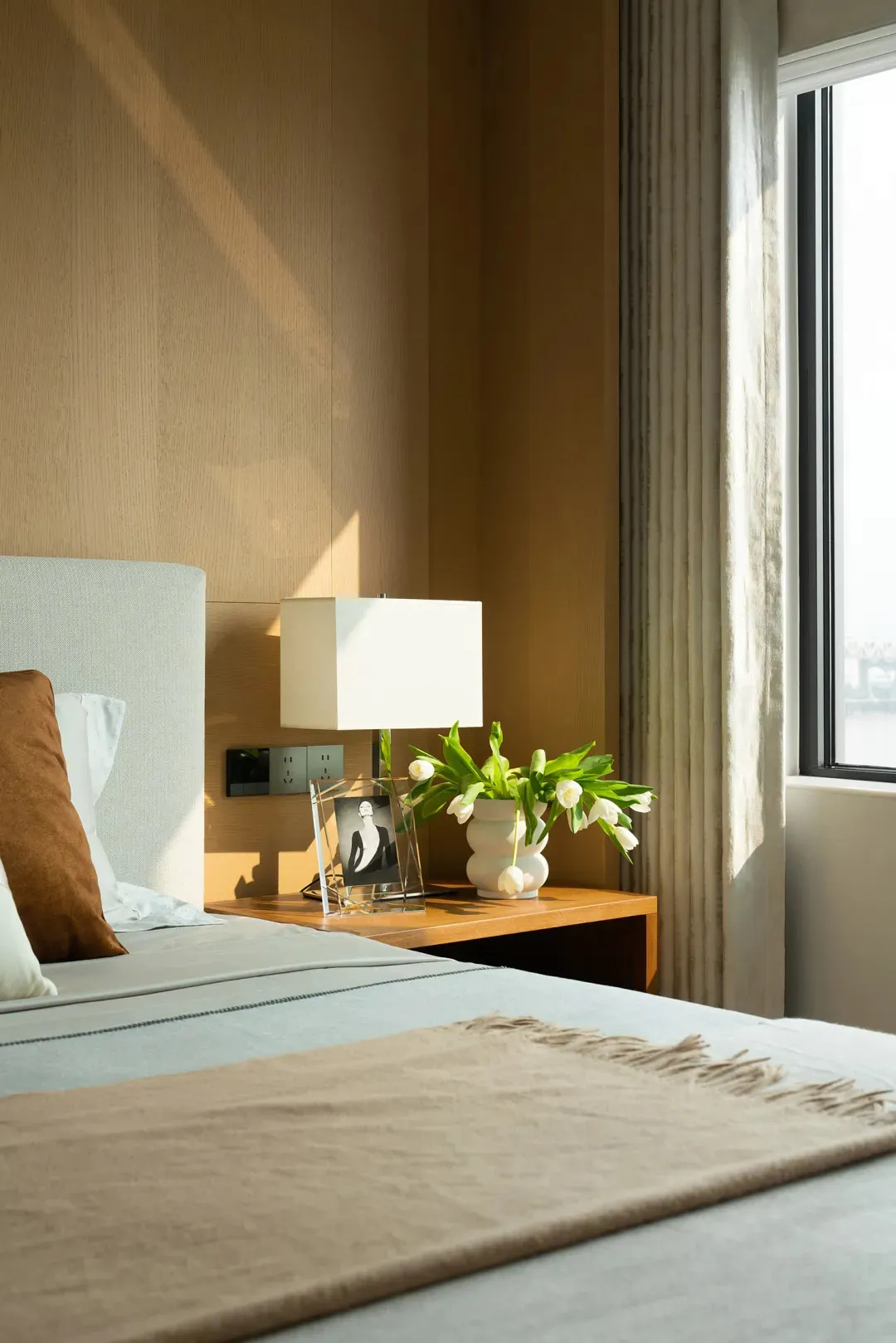 A modern minimalist bedroom featuring a wooden bed frame, fabric bedding, and a glass lamp and vase with white tulips.