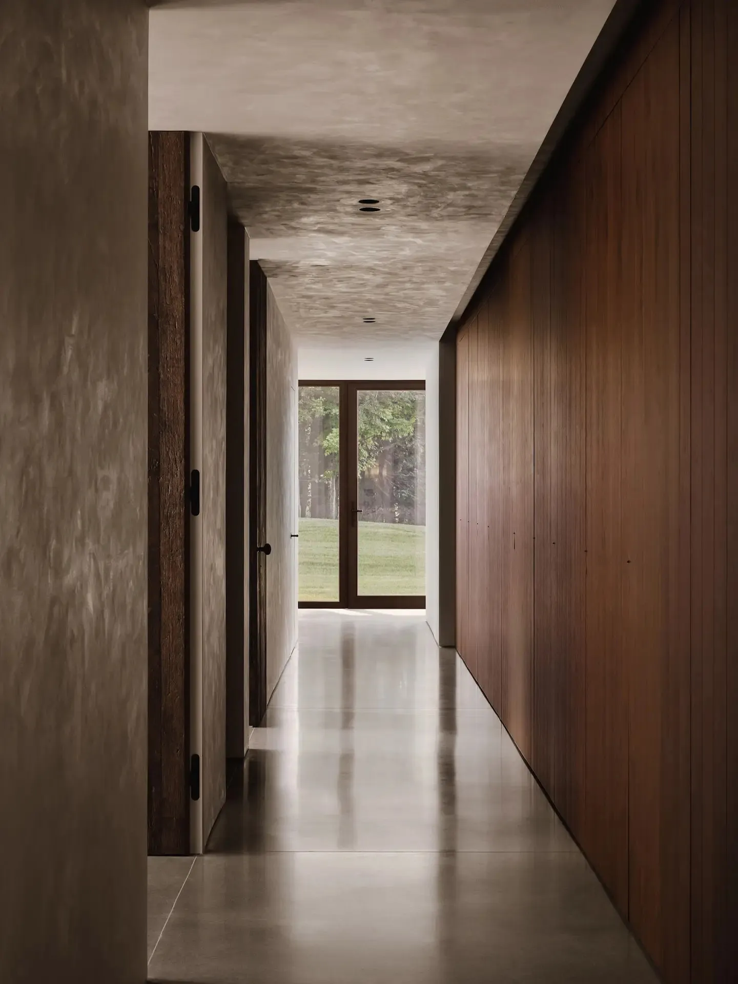 A modern minimalist hallway with concrete flooring and walls, featuring wooden doors and a glass door leading to a green outd
