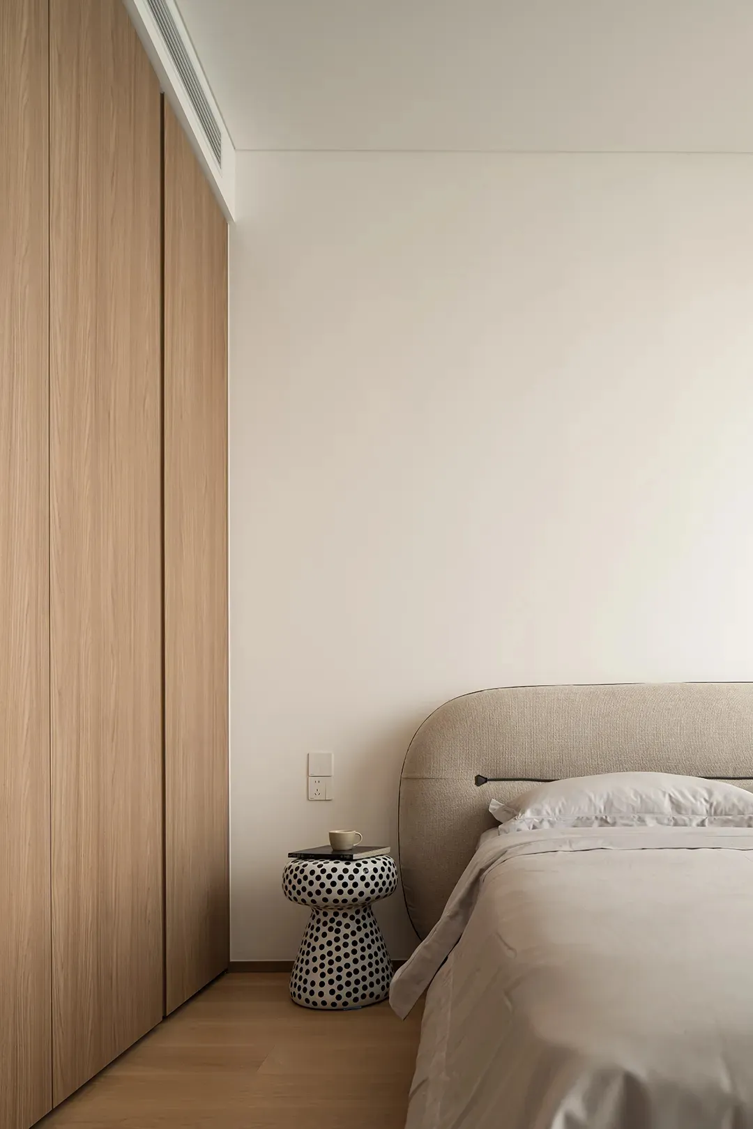 Minimalist bedroom with light wood flooring, a beige upholstered bed, and a patterned side table with a mug and book.