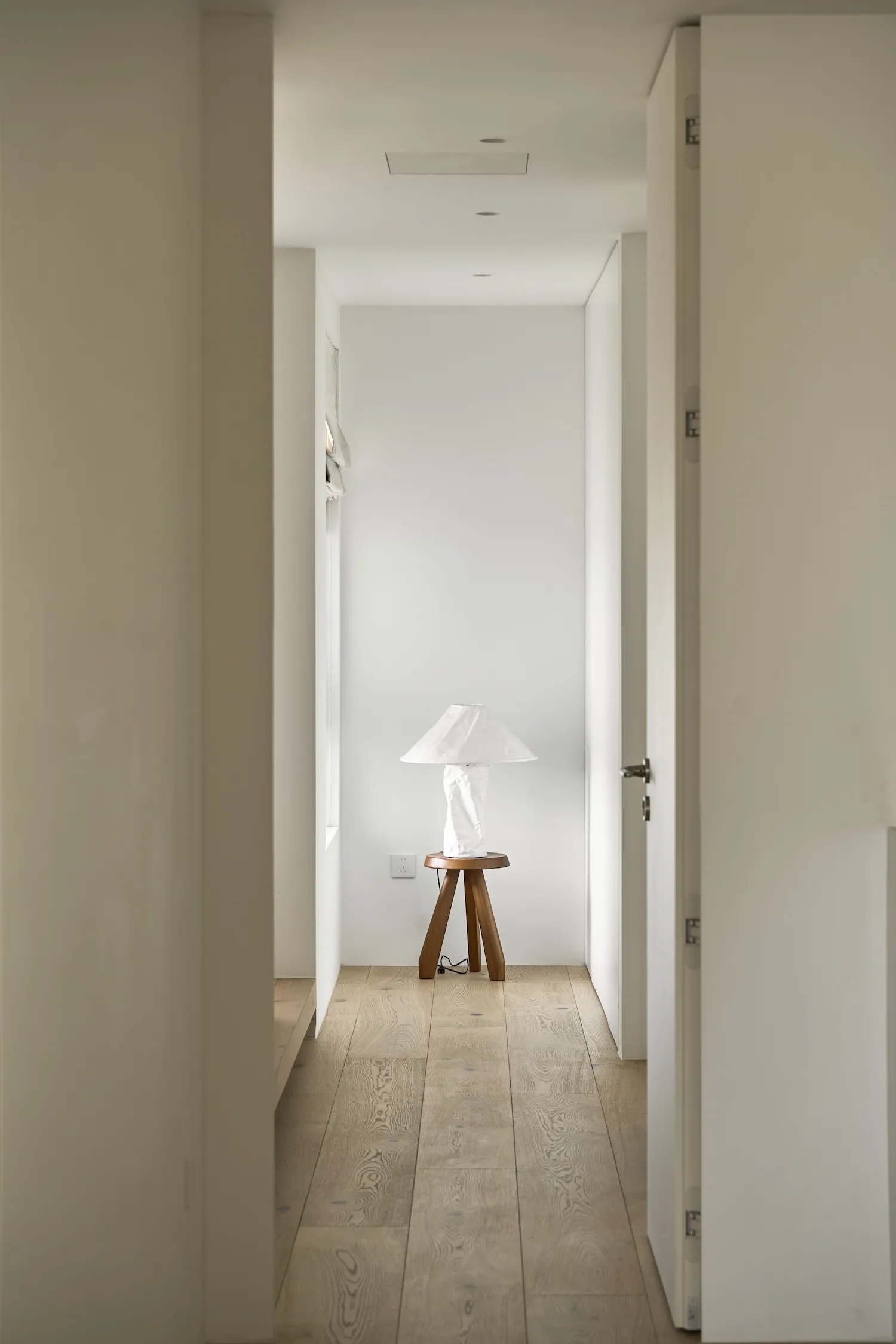A minimalist corridor with white walls, wooden flooring and a small, wooden tripod table with a white lampshade