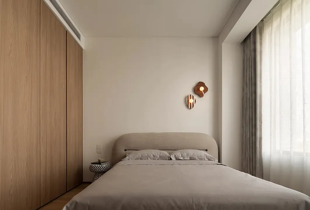 A minimalist bedroom featuring a bed with a neutral-colored headboard and bedspread, along with a simple nightstand and decor