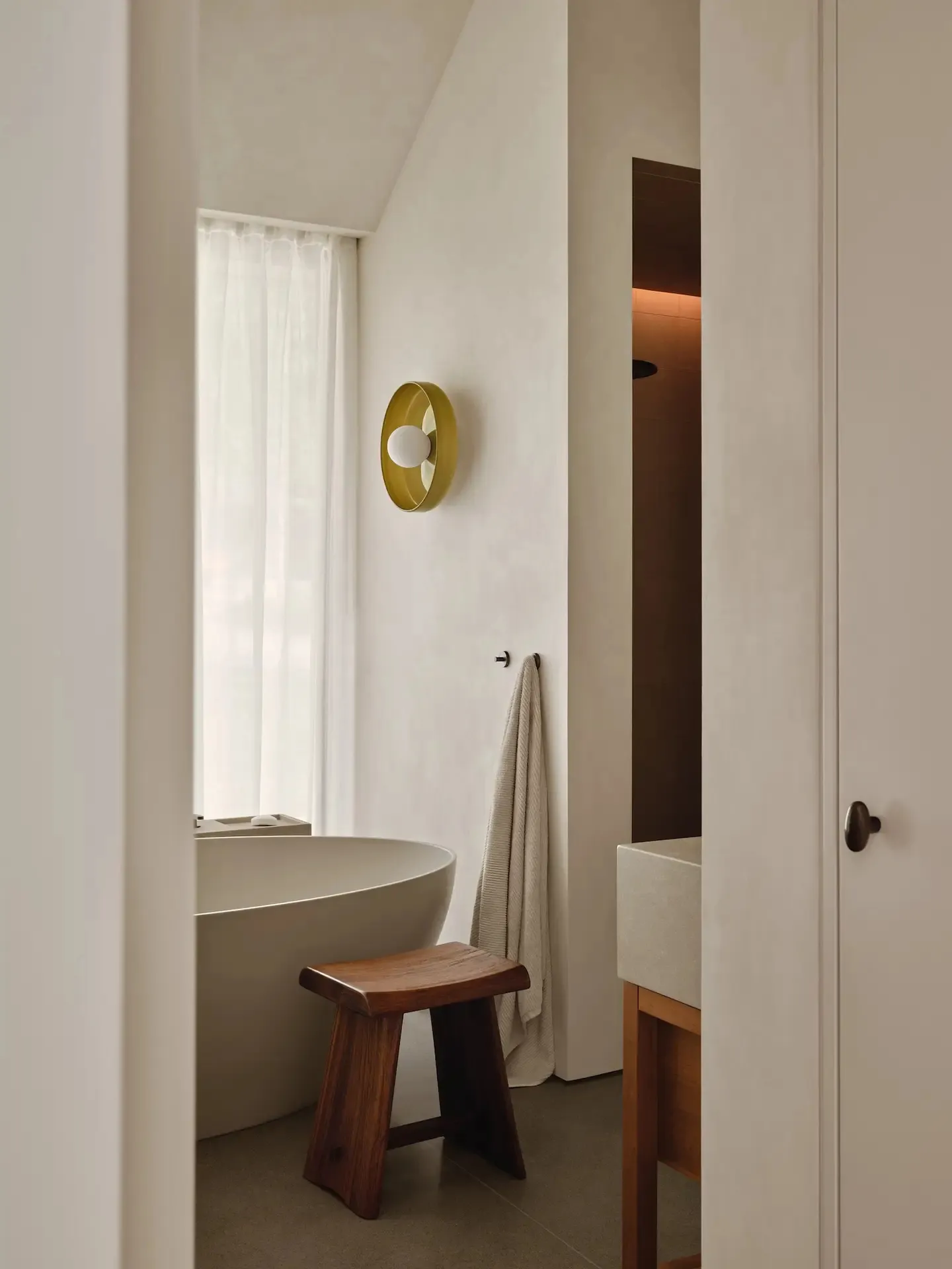 A modern bathroom featuring concrete walls and a minimalist design, with a freestanding bathtub, wooden stool, and golden cir