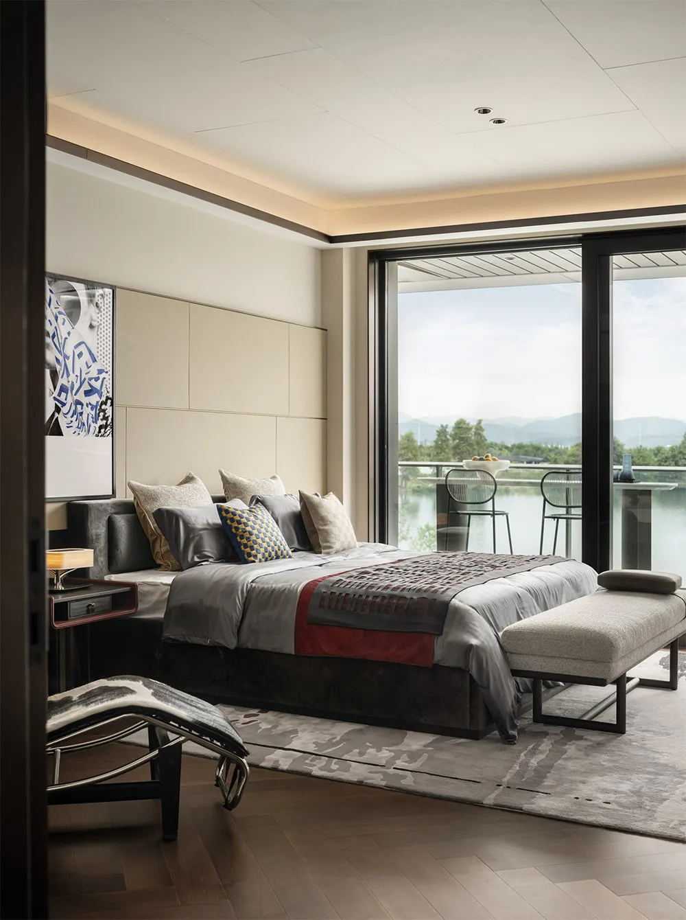 A modern bedroom with fabric bedding, a wooden floor, and a glass door overlooking a scenic landscape.