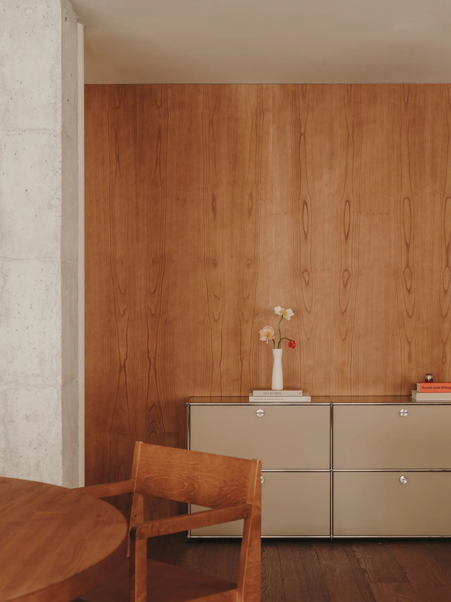 A minimalist interior design featuring a wooden chair, table, and cabinet with a vase of flowers and books on top.