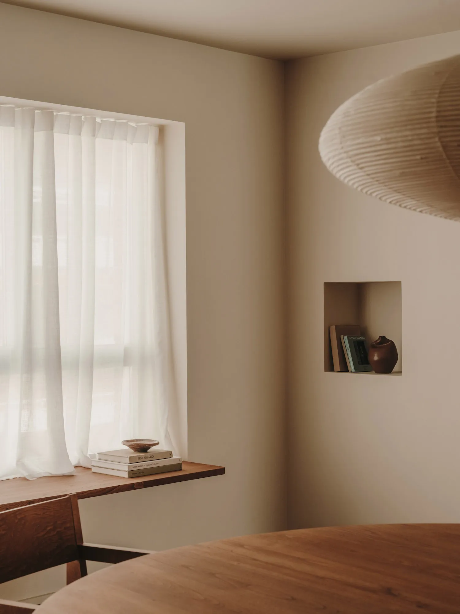 A minimalist interior with a wooden shelf and white sheer curtains, featuring a suspended lamp and a niche with books and a v
