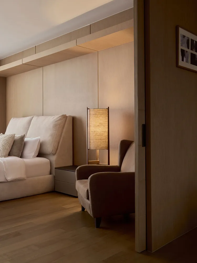 A modern minimalist bedroom featuring a wooden floor, upholstered furniture, and a floor lamp with a woven shade.