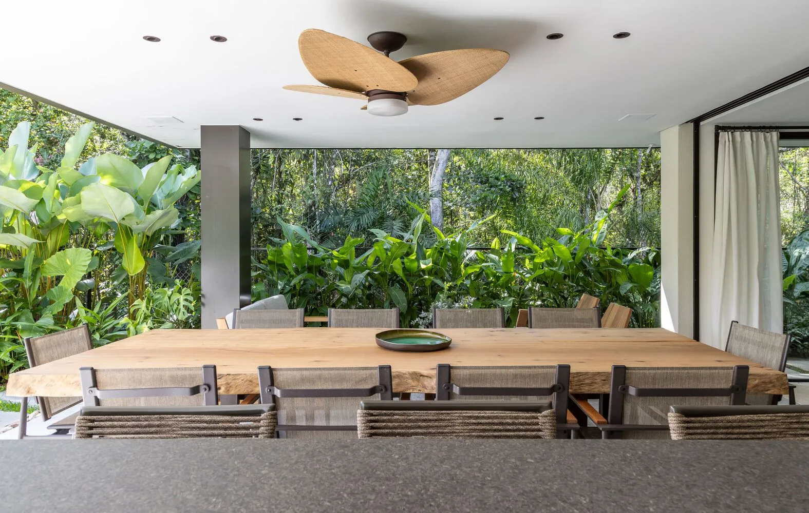 Modern dining room with a wooden table and chairs surrounded by greenery and large windows.