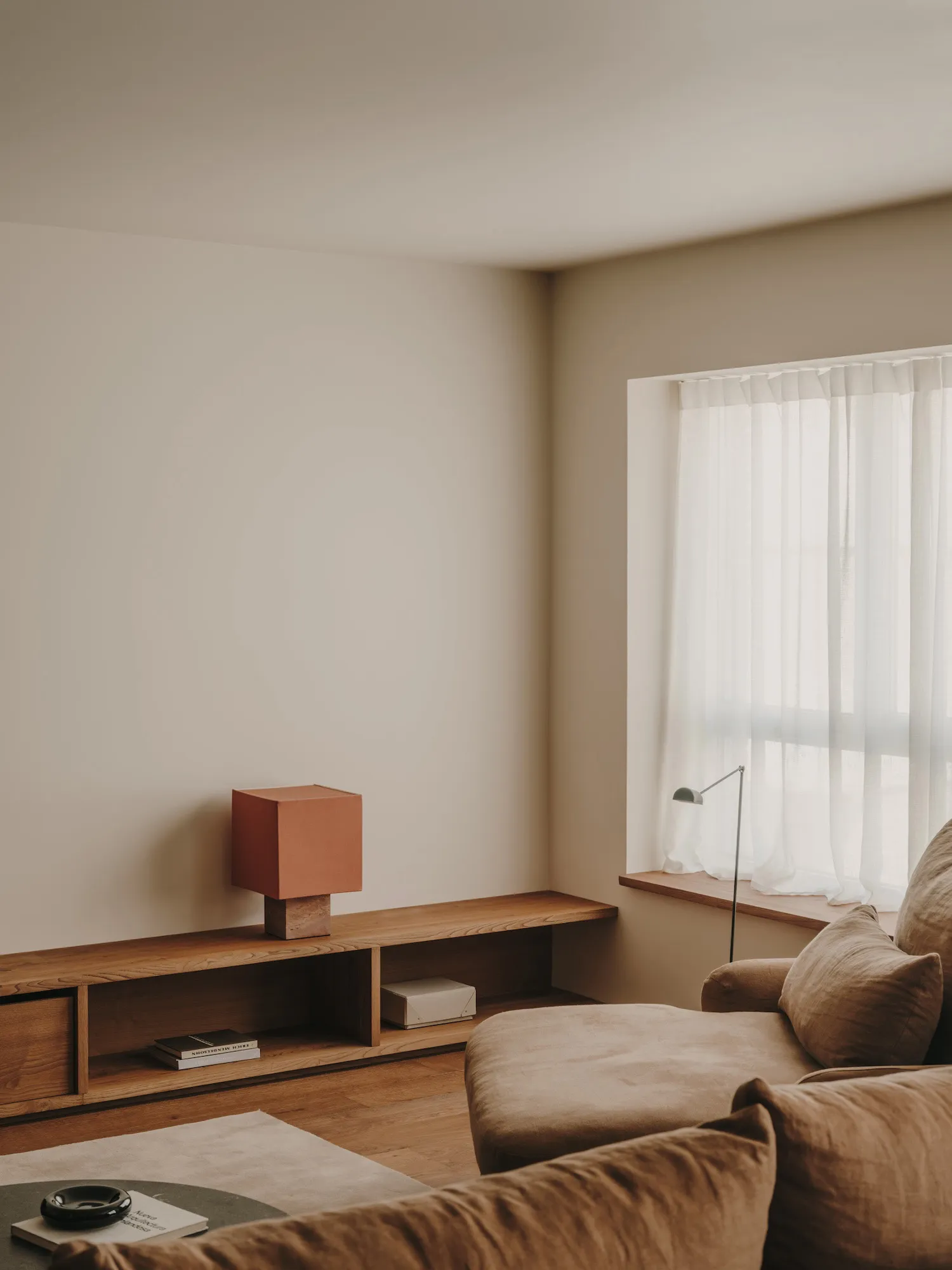 A modern minimalist living room featuring a wooden sofa and table with a red square lamp on a wooden shelf, next to a window