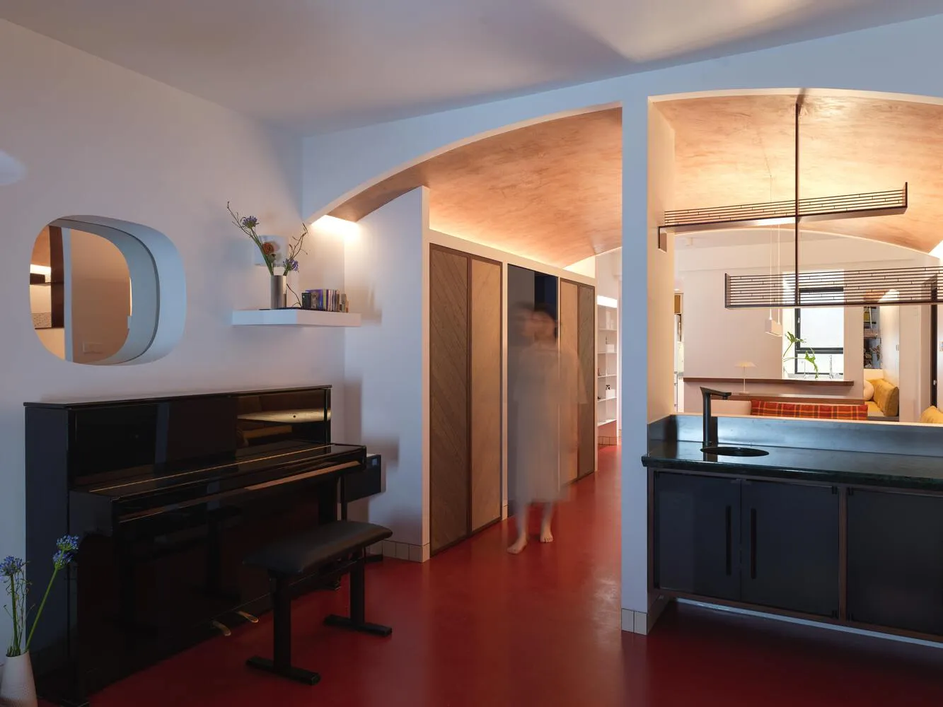 A modern living space featuring an open-plan design with a piano, kitchen area, and bookshelf.