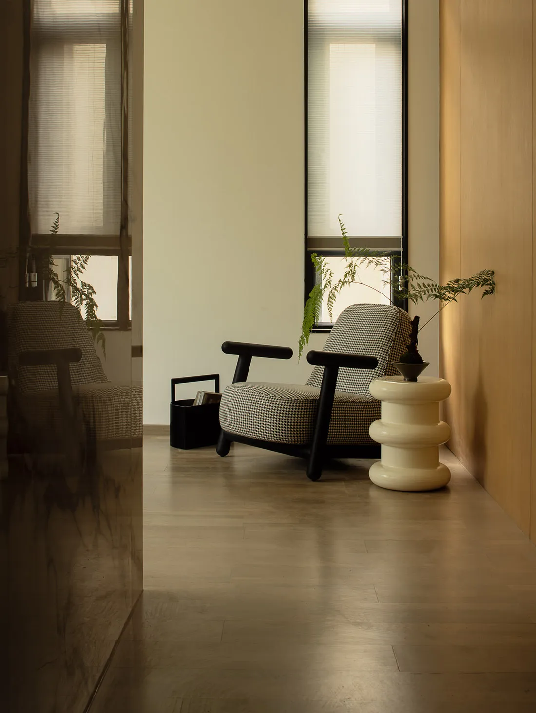 A minimalist monochrome living room with wooden floors and white walls, featuring a checkered armchair, side table, and plant