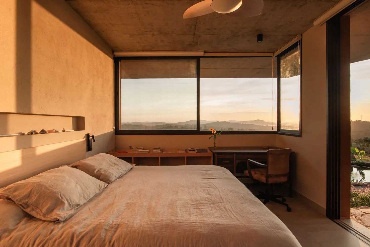 A modern minimalist bedroom with concrete walls, large windows, and a view of the mountains at sunset.