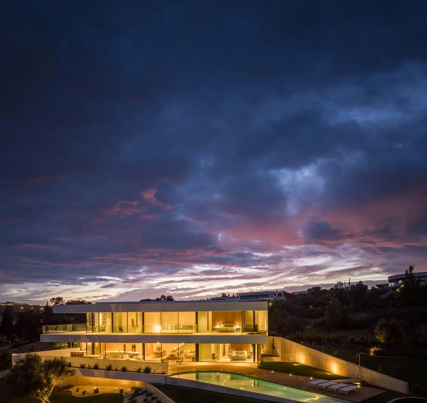 Modern residential exterior with a glass and concrete facade illuminated during the evening with the sky in the background.