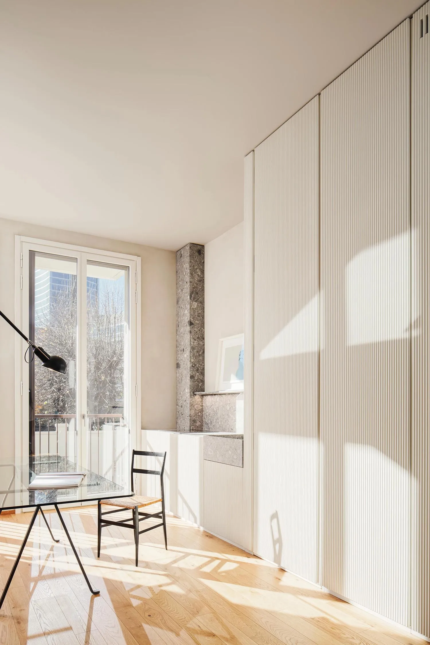 A modern home office featuring wooden flooring and a glass table with a black lamp and a wooden chair.