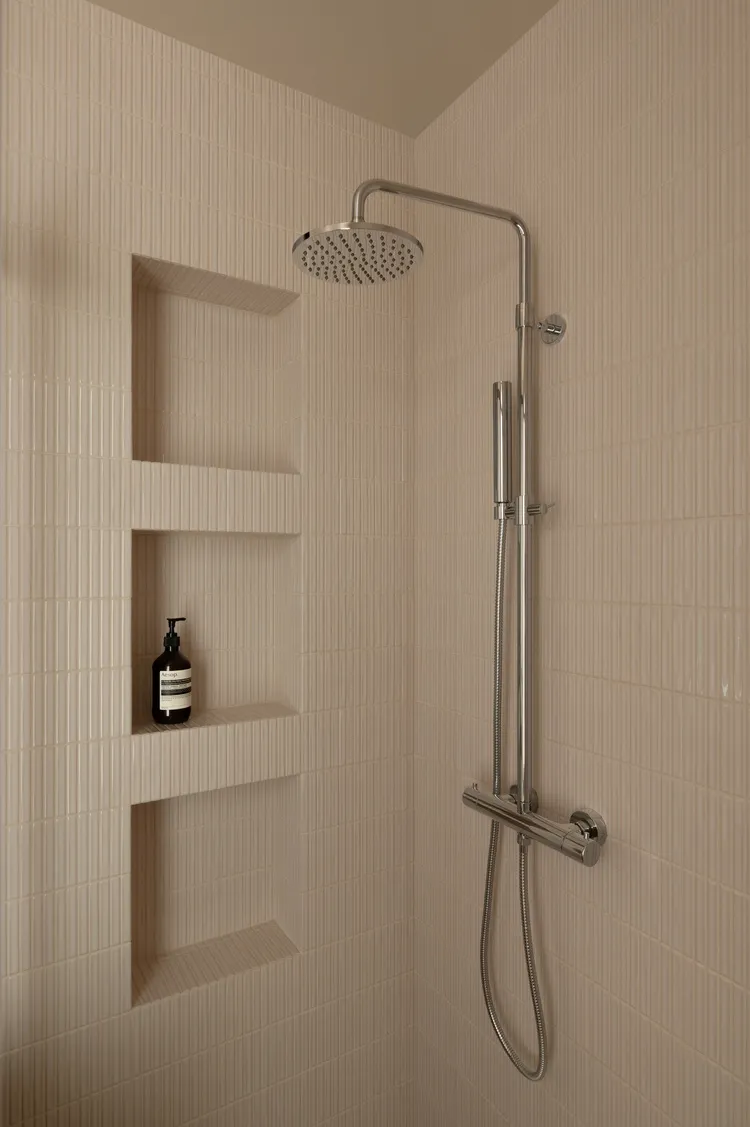 A modern minimalist shower with textured beige tiles, featuring a fixed showerhead and a handheld showerhead mounted on a chr