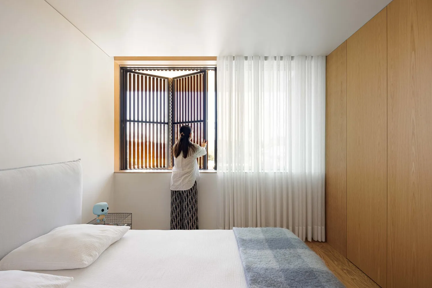 A person standing in front of a window with wooden shutters and blinds in a modern minimalist bedroom setting.