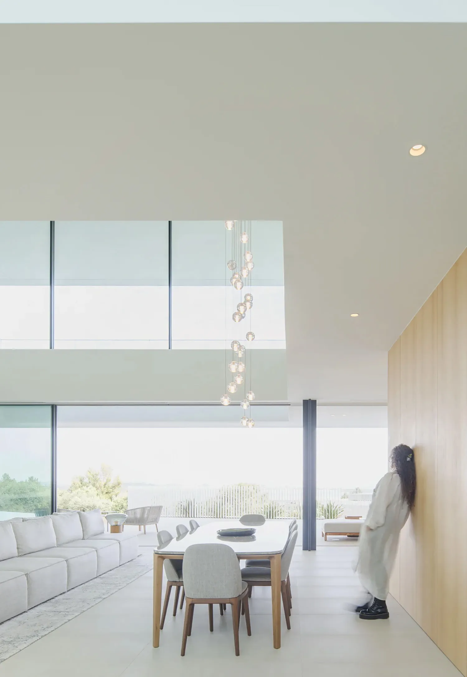 A minimalist living room with large windows and concrete walls, featuring a white sectional sofa, wooden dining table, and mo
