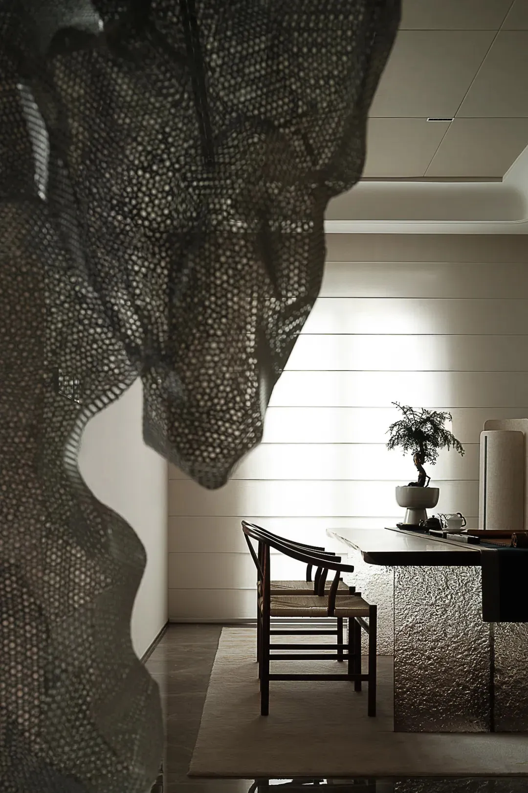 A modern dining room with a textured fabric screen, wooden chairs, and a bonsai tree on a polished dining table.