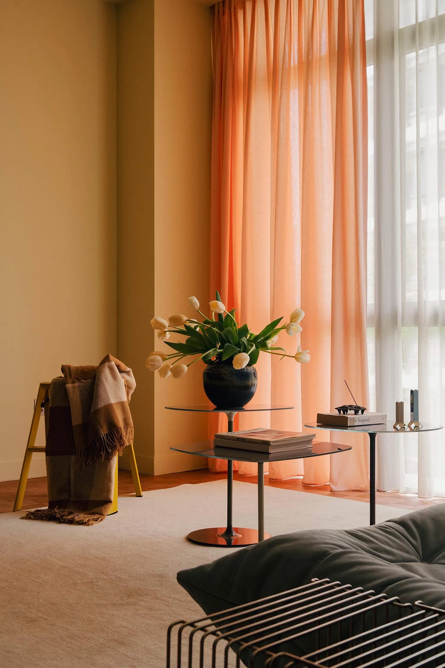 A cozy living room with a gray sofa, orange curtains, and white tulips in a vase on a glass table.