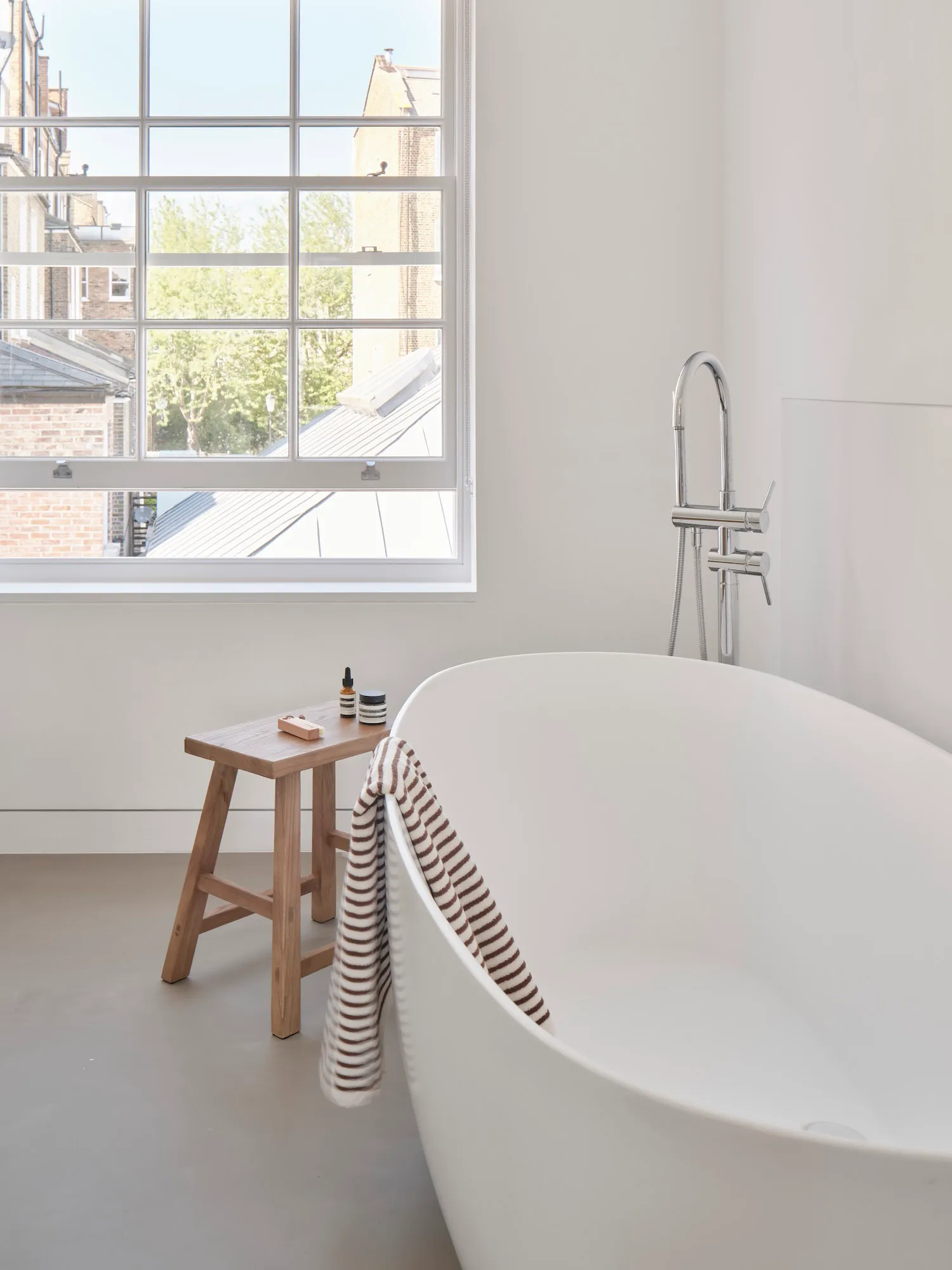 A modern white bathroom with a freestanding ceramic tub and a stool with bottles and a sponge on top.