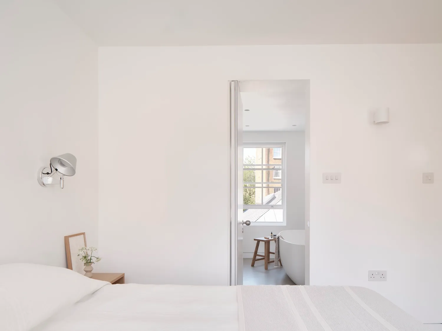 A minimalist bedroom with white walls, bed, and bathroom in the background, featuring a white bathtub and wooden stool.