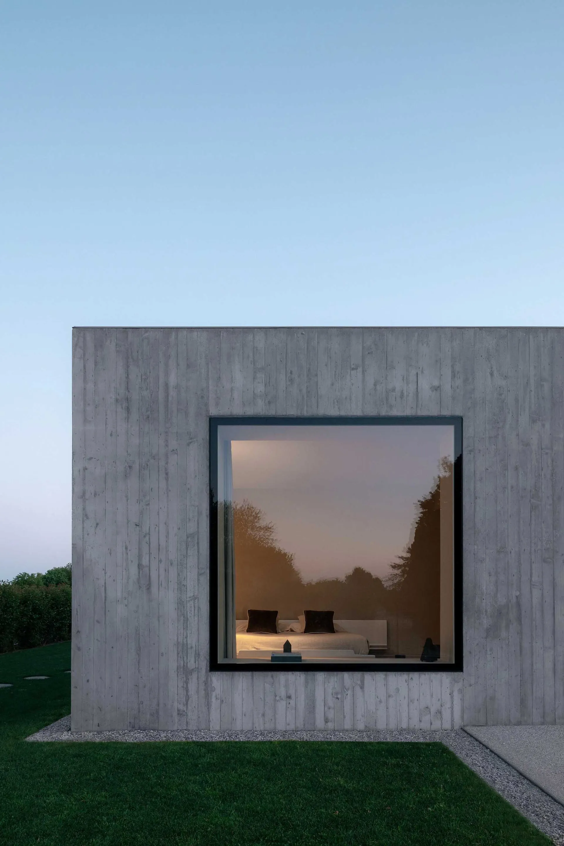 Modern, minimalist concrete box house with a large window reflecting trees and the interior bedroom.