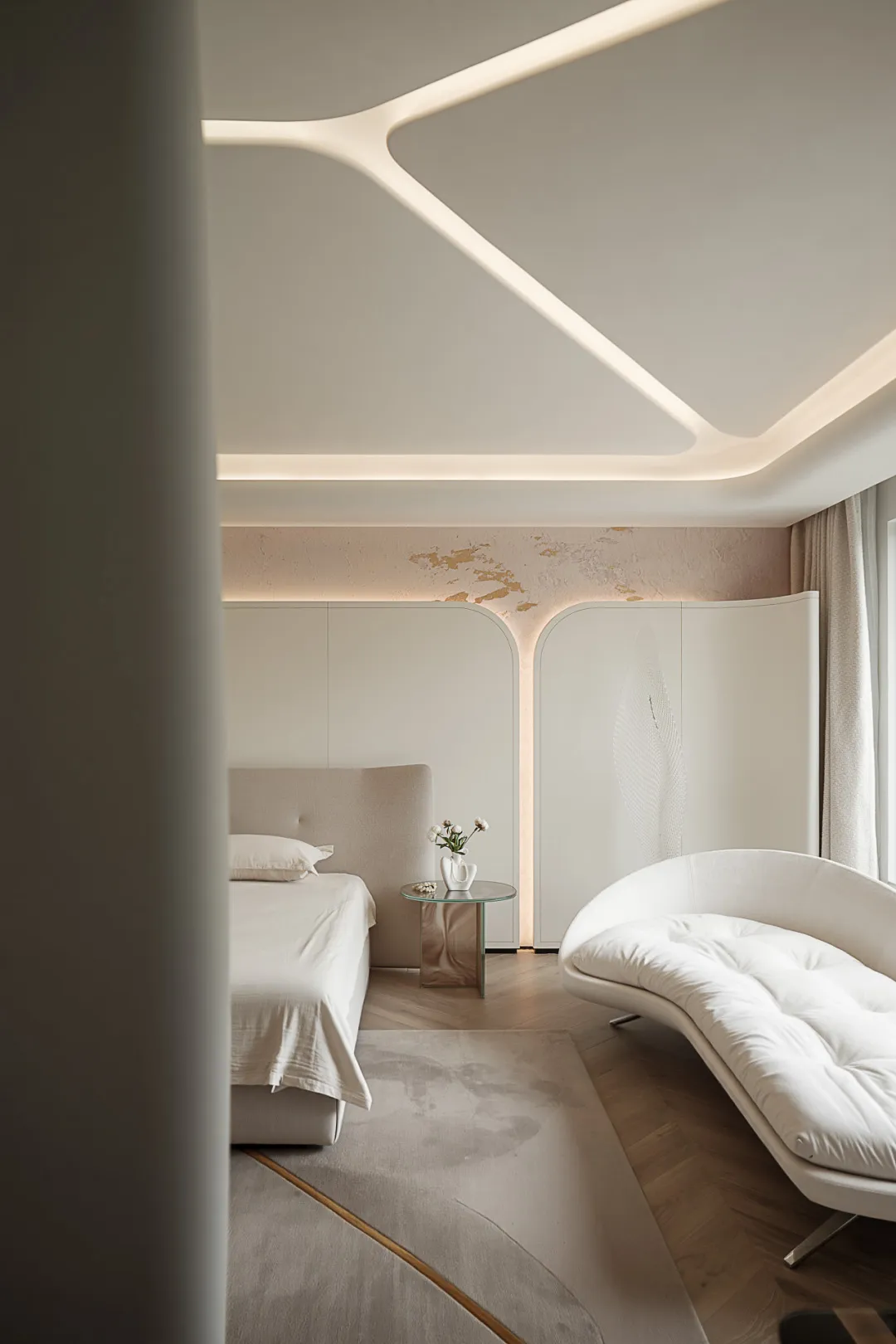 Minimalist bedroom with a beige bed and a curved white sofa, featuring a glass bedside table with a vase of flowers.