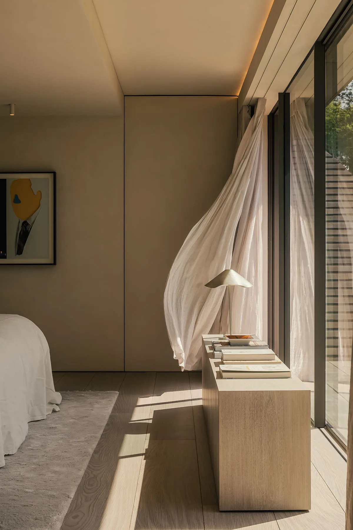 Minimalist bedroom interior with a bed, a window with sheer curtains, and a nightstand with a lamp and books.