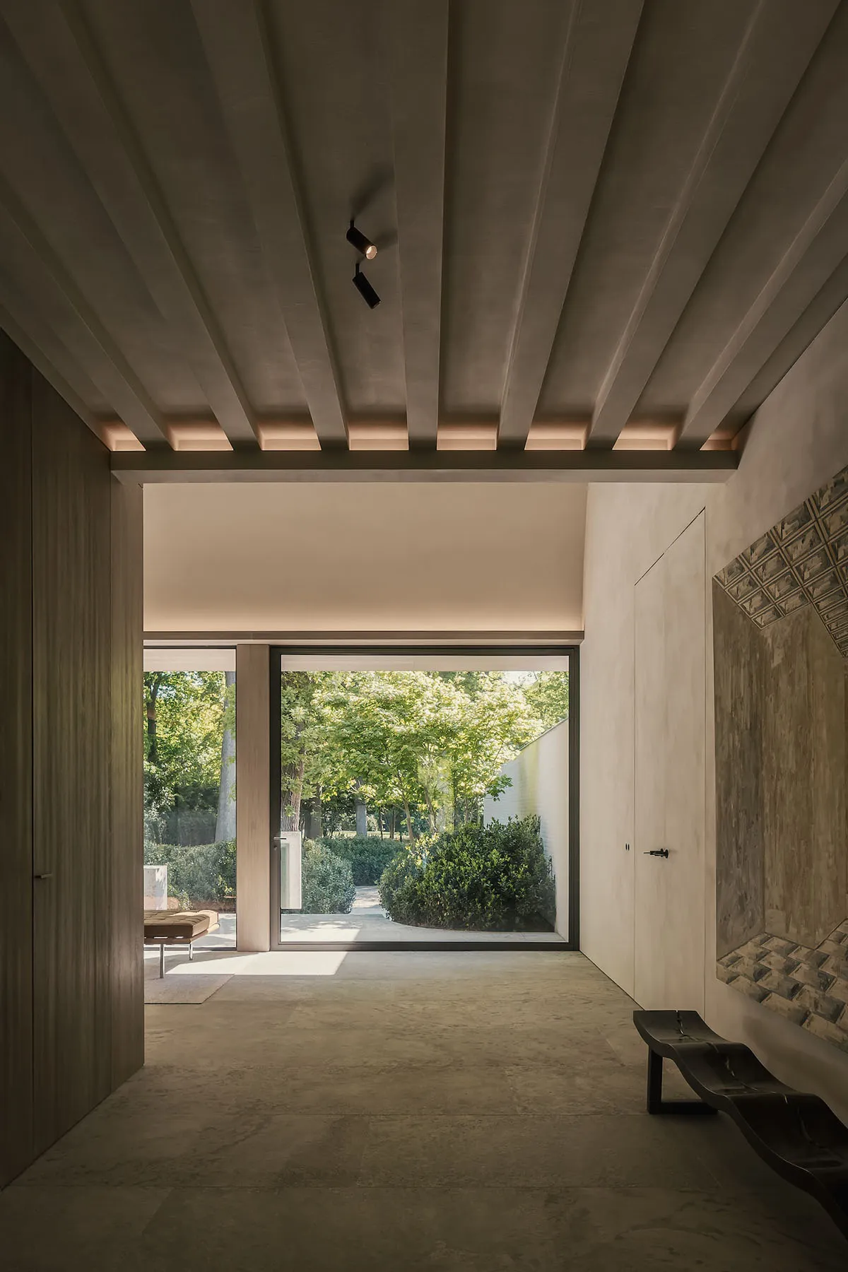 Interior design of a minimalist living room with concrete flooring and walls, featuring a large window with greenery outside.