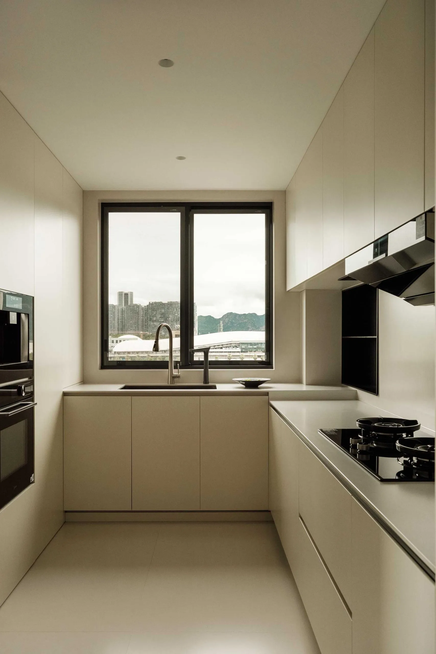 A modern minimalist white kitchen with a large window providing a view of the cityscape and green mountains.
